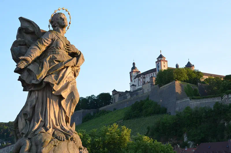 Brückenfigur der Franconia auf der Alten Mainbrücke in Würzburg mit der Festung Marienberg im warmen Abendlicht