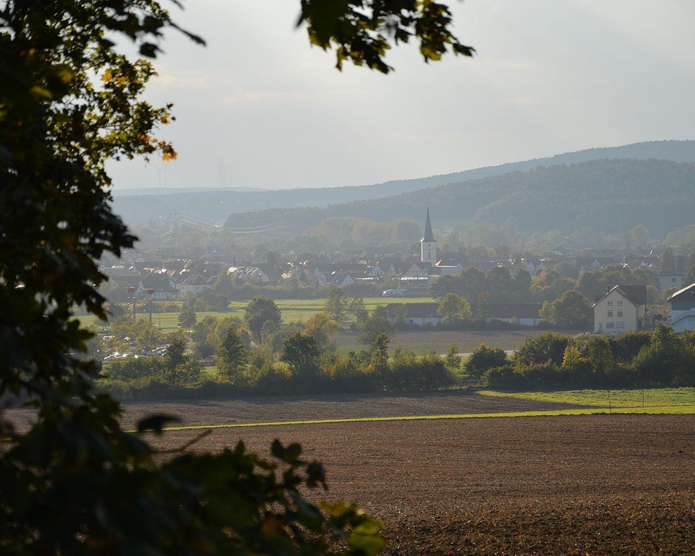 Blick über Felder auf ein Dorf mit Kirchturm und bewaldeten Hügeln